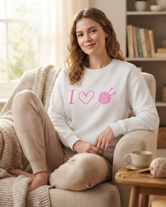 A woman sits in a cozy living room on an armchair, wearing a white crewneck sweatshirt with a pink graphic that reads 'I [heart] knitting yarn' and beige knit pants.
