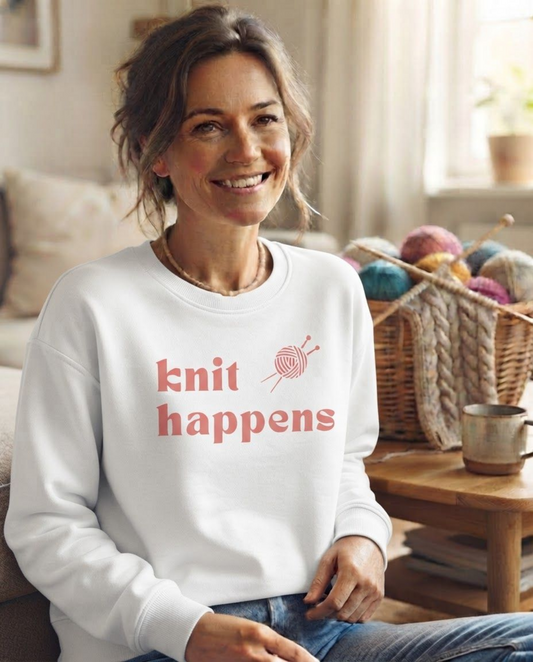 A smiling woman sits in a cozy living room wearing a white crewneck sweatshirt with the phrase 'knit happens' and a small knitting needle graphic in coral pink. A basket of colorful yarn and a wooden mug are visible in the background.