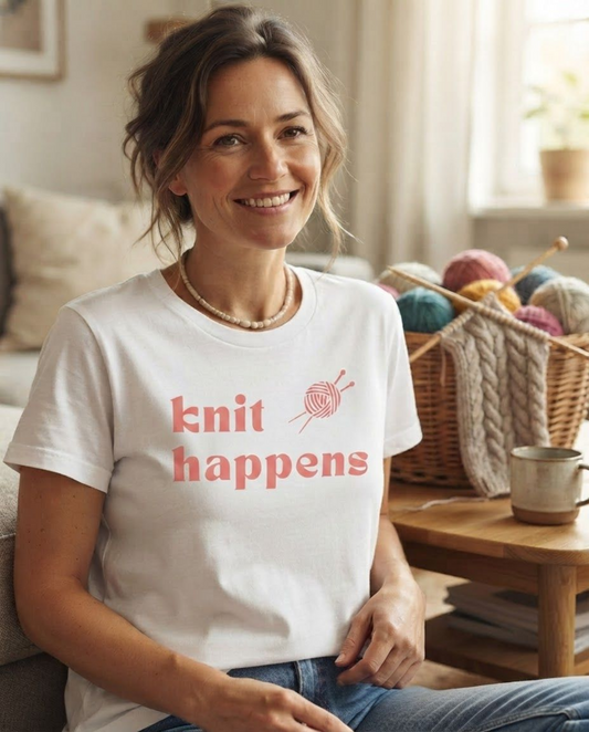 A smiling woman sits in a cozy living room wearing a white graphic T-shirt with the words 'knit happens' in coral pink and a small knitting needle and ball graphic. A basket of colorful yarn and a mug sit on a wooden table in the background.