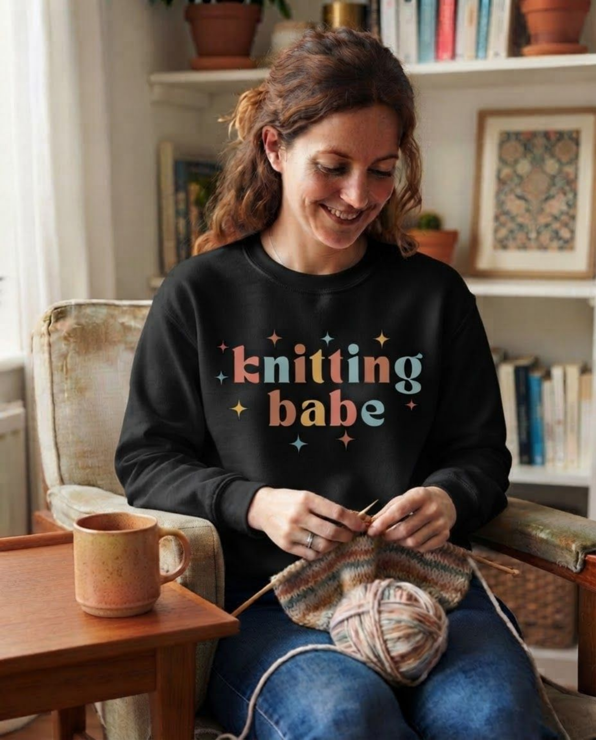 A woman sitting in a cozy living room setting, knitting with wooden needles and a ball of yarn while wearing a black sweatshirt with the text 'knitting babe' in colorful pastel letters. A wooden mug sits on the side table next to her.