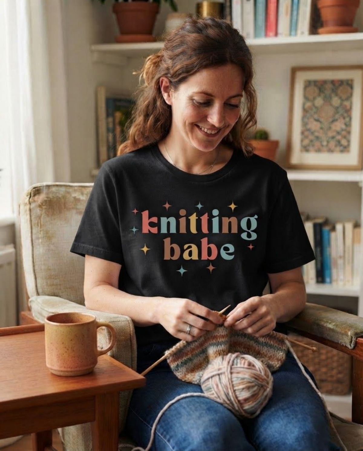 A woman sitting in a cozy room, knitting with yarn while wearing a black graphic t-shirt that reads 'knitting babe' in colorful letters. A wooden mug sits on the table next to her.