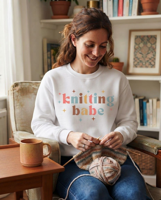 A woman sitting in a cozy armchair, knitting with colorful yarn while wearing a white sweatshirt with the text 'knitting babe' printed on the front in pastel colors. A wooden mug sits on the table next to her.