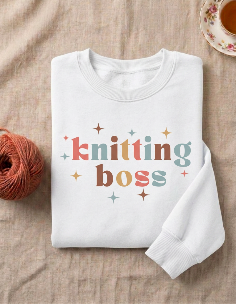 A folded white crewneck sweatshirt with the words 'knitting boss' printed in a retro multicolor font with small star accents, styled on a beige linen fabric with a ball of orange yarn and a teacup in the background.