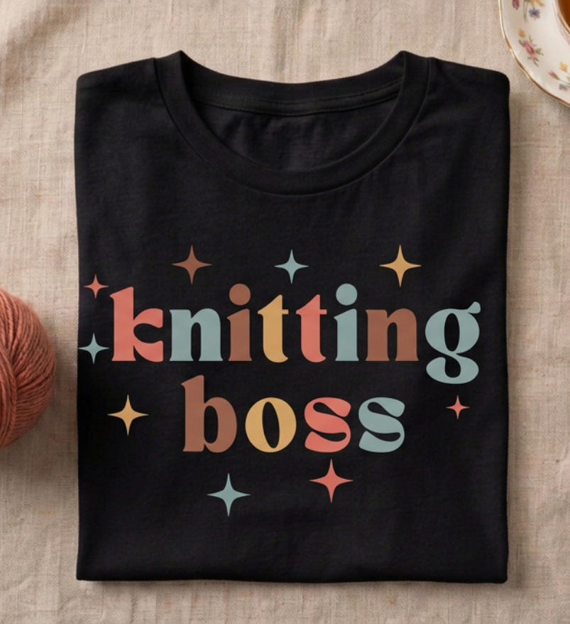 A folded black t-shirt with a retro-style colorful graphic that reads 'knitting boss' in pastel orange, yellow, and blue-green lettering, surrounded by small star accents. The shirt is laid flat on a beige linen surface next to a ball of brown yarn.