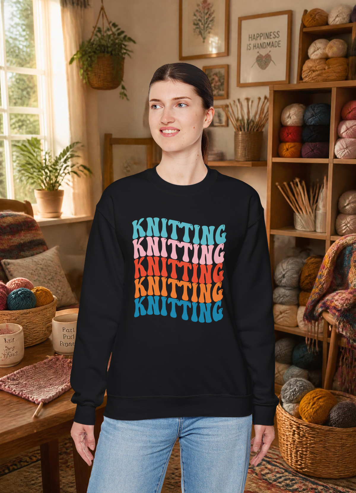 A woman wearing a black crewneck sweatshirt with a colorful retro-style stacked 'KNITTING' graphic on the front, standing in a cozy yarn shop interior.