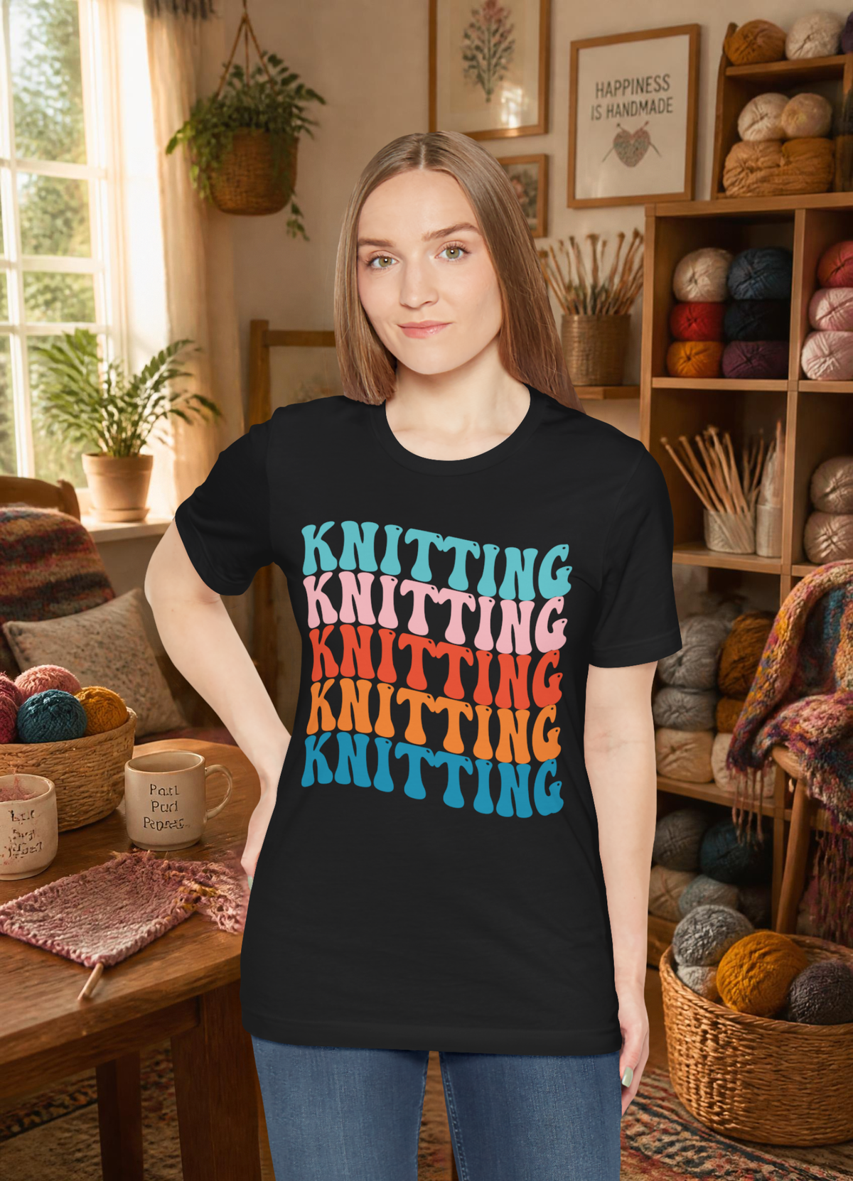 A woman modeling a black graphic t-shirt with the word 'KNITTING' repeated in a colorful retro gradient. The background is a cozy, warm room filled with shelves of yarn, knitting needles, and knitted blankets.