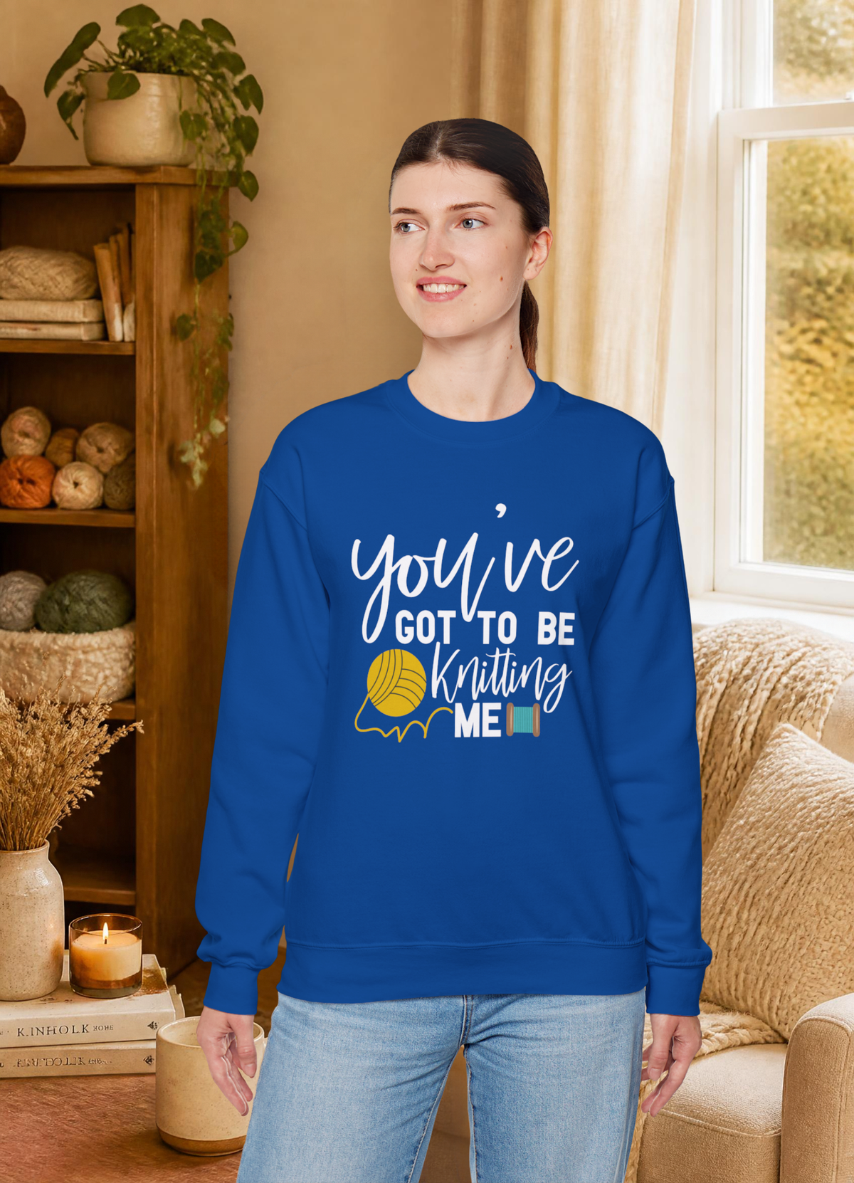 A woman wearing a royal blue crewneck sweatshirt with a white graphic that reads 'you've GOT TO BE Knitting ME' alongside a yellow yarn ball illustration, standing in a cozy, warm-toned living room.