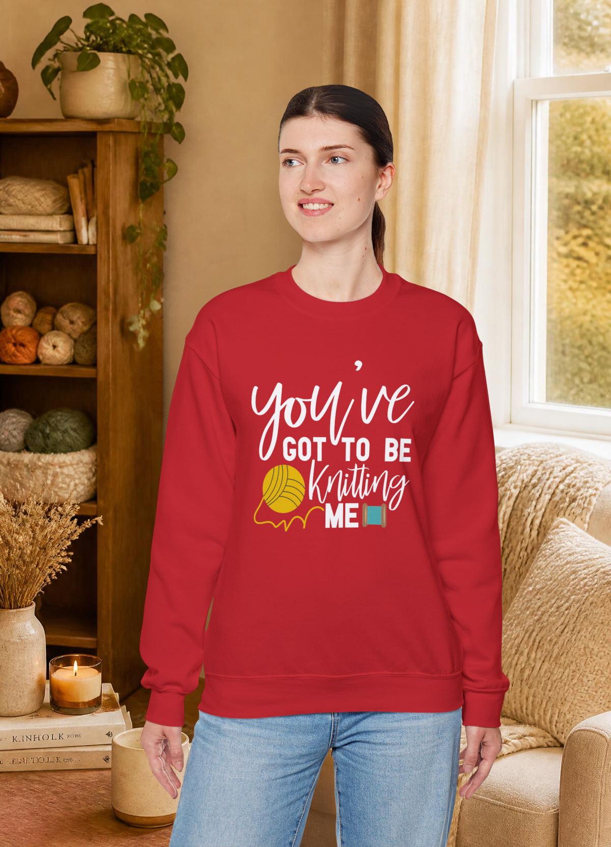 A woman wearing a red crewneck sweatshirt with a white graphic that reads 'you've GOT TO BE Knitting ME' with a yellow yarn ball illustration, standing in a cozy, warm-toned room with a bookshelf and window in the background.