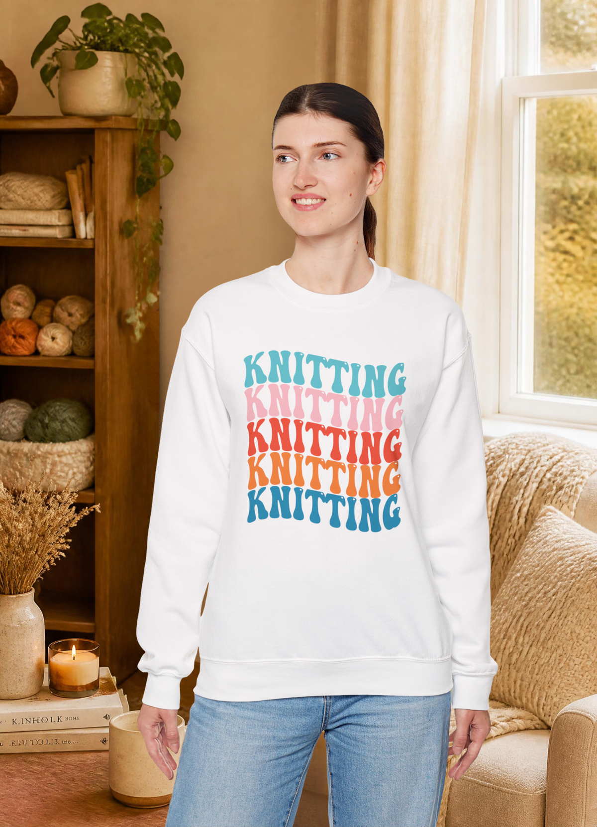 A woman wearing a white crewneck sweatshirt with a colorful stacked 'KNITTING' graphic, paired with light blue jeans, standing in a cozy, warm-toned room with a bookshelf and window.