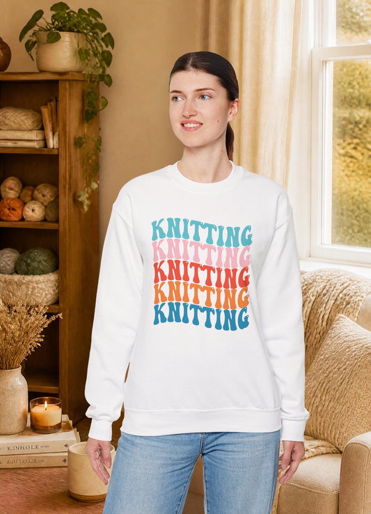 A woman wearing a white crewneck sweatshirt with a colorful stacked 'KNITTING' graphic, paired with light blue jeans, standing in a cozy, warm-toned room with a bookshelf and window.