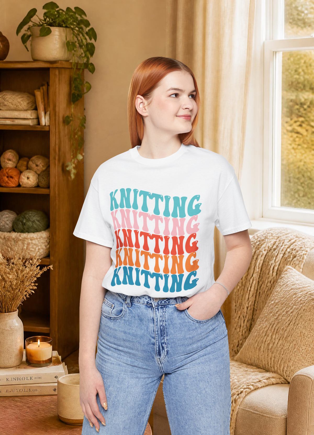 A woman with red hair stands in a cozy room wearing a white graphic T-shirt with the word 'KNITTING' repeated in stacked colorful letters (teal, pink, orange, yellow) and light blue jeans.