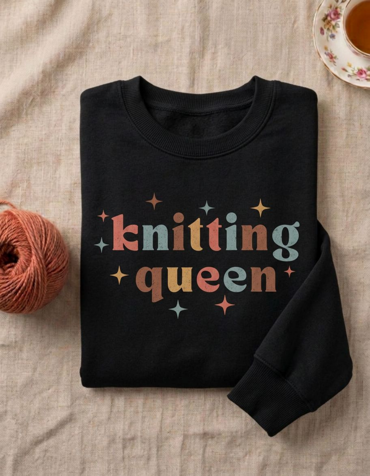 A folded black crewneck sweatshirt with the words 'knitting queen' in retro multicolor letters, styled on a beige linen fabric with a ball of orange yarn and a cup of tea in the background.