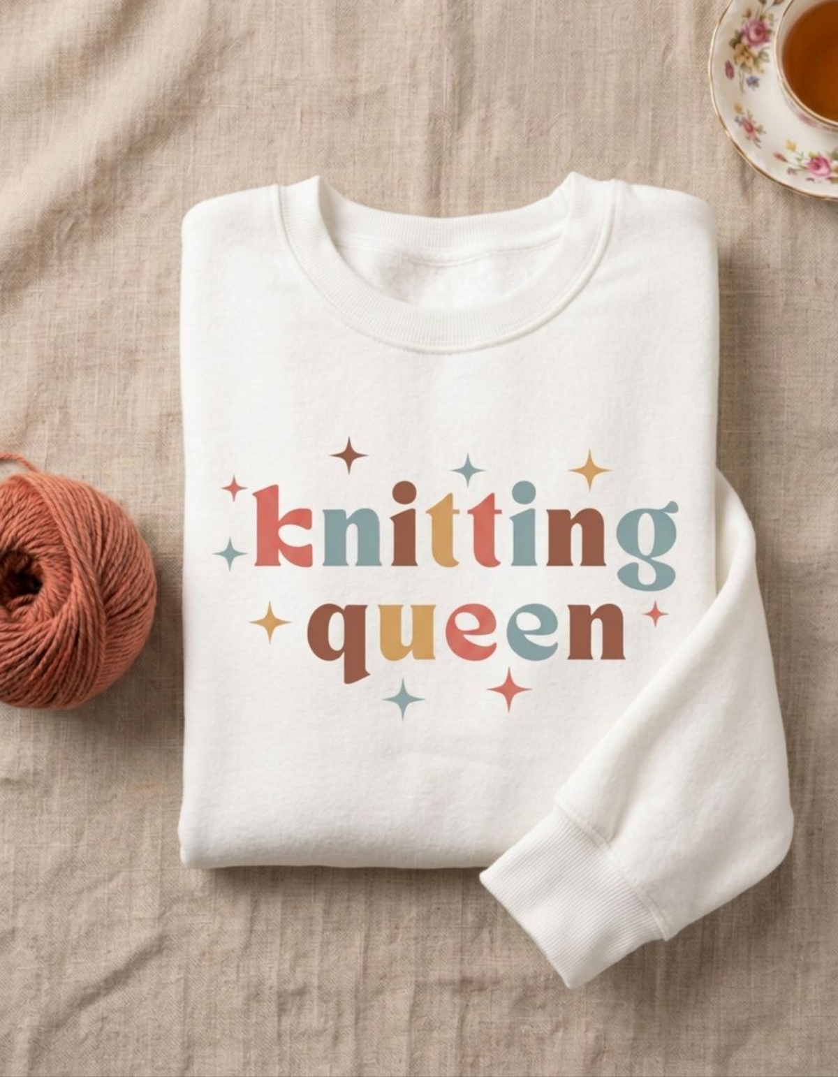 A folded white crewneck sweatshirt with the words 'knitting queen' in colorful retro letters and small star accents, styled on a beige linen surface with a ball of orange yarn and a teacup in the background.