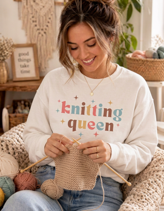 A woman smiling while knitting, wearing a cream sweatshirt with the words 'knitting queen' printed in colorful letters. She is sitting in a cozy room with yarn balls and knitted blankets.