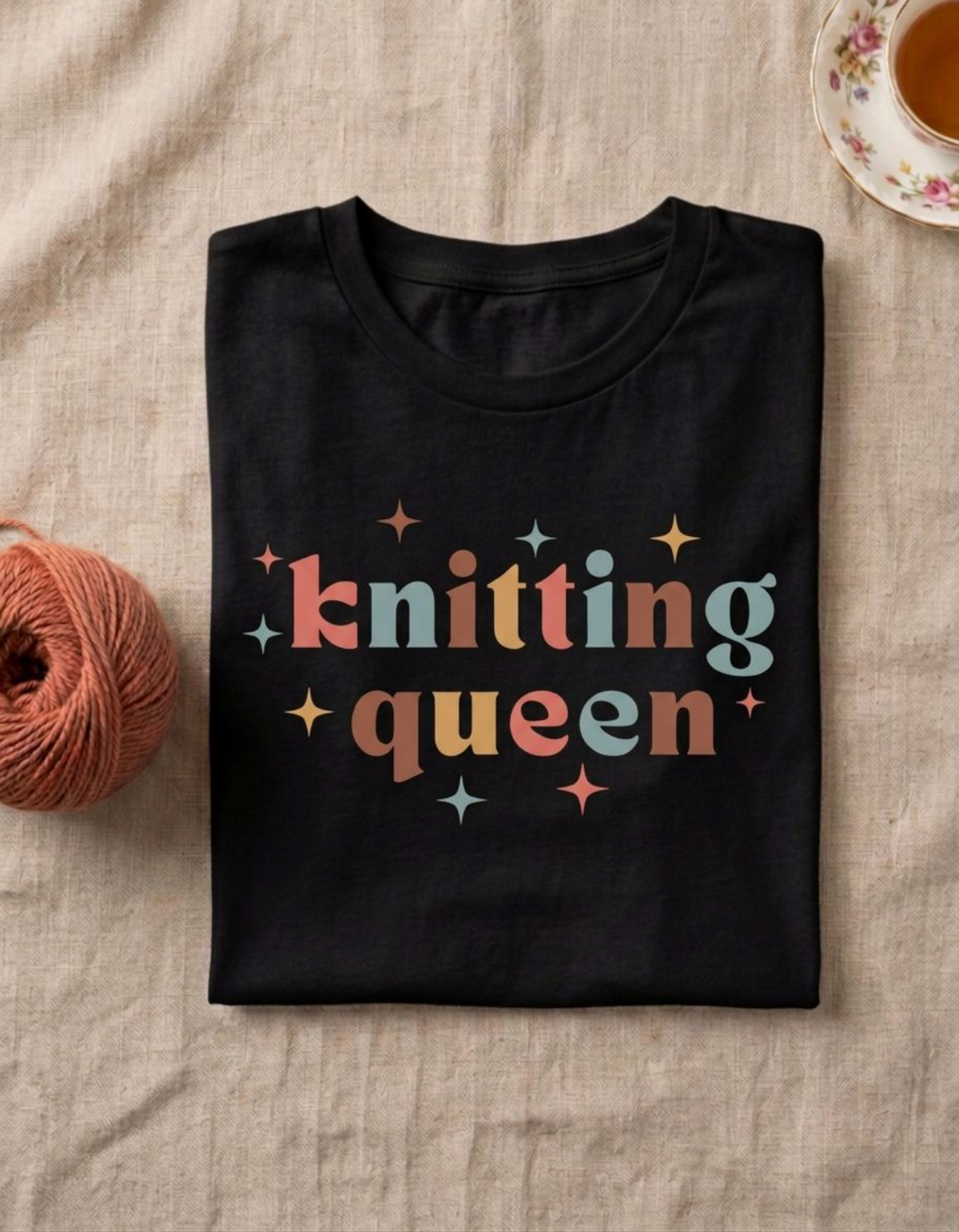 A folded black t-shirt with a colorful retro-style graphic reading 'knitting queen' surrounded by small stars, styled on a beige linen fabric with a ball of orange-brown yarn on the left and a floral teacup on the top right.