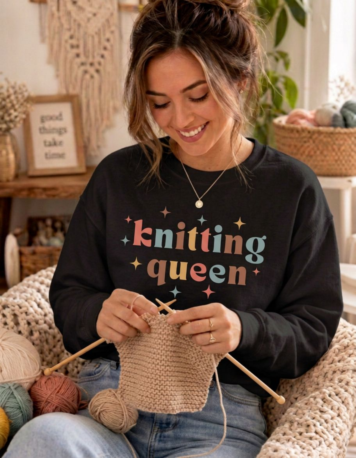 A woman smiling while knitting, wearing a black sweatshirt with the words 'knitting queen' printed in colorful letters. She is seated in a cozy room with yarn balls and knitted blankets.
