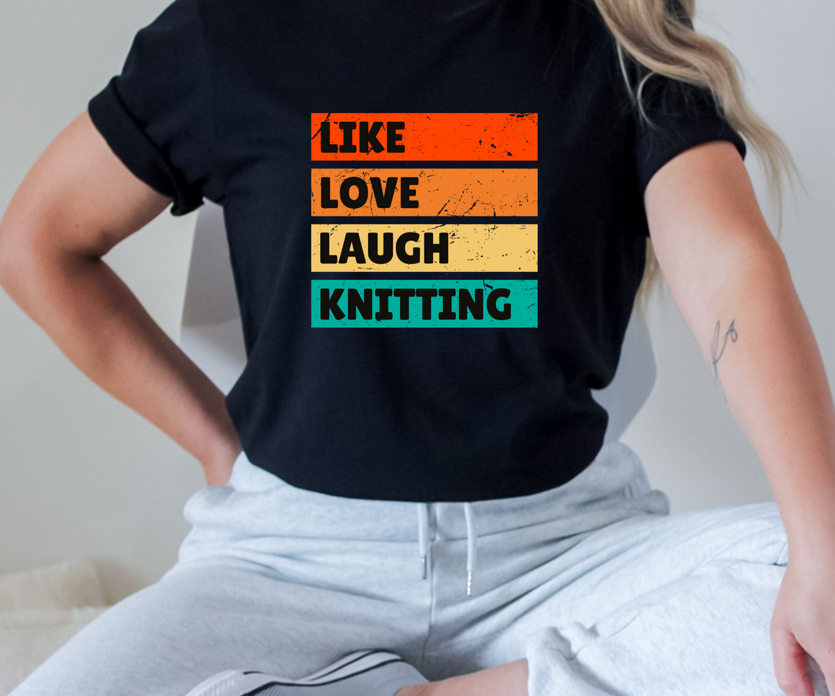 A person wearing a black short-sleeve T-shirt with a retro-style graphic that reads 'LIKE LOVE LAUGH KNOTTING' in stacked horizontal stripes of orange, yellow, beige, and teal. The person is also wearing light gray sweatpants and is seated indoors.