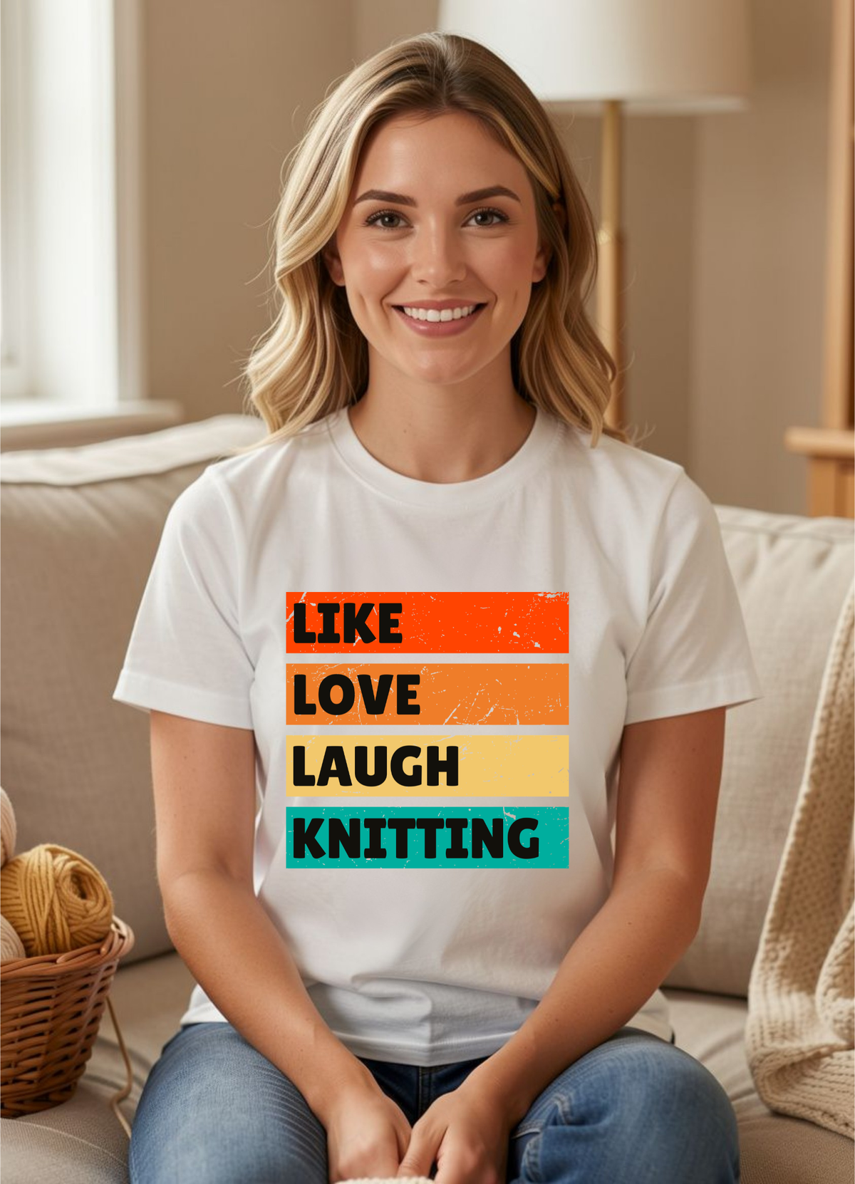 A smiling blonde woman sits on a beige couch wearing a white graphic T-shirt with the phrase 'LIKE LOVE LAUGH KNITTING' printed in stacked colorful blocks. The background shows a softly lit living room with a lamp, basket, and knit blanket.