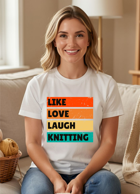 A smiling blonde woman sits on a beige couch wearing a white graphic T-shirt with the phrase 'LIKE LOVE LAUGH KNITTING' printed in stacked colorful blocks. The background shows a softly lit living room with a lamp, basket, and knit blanket.