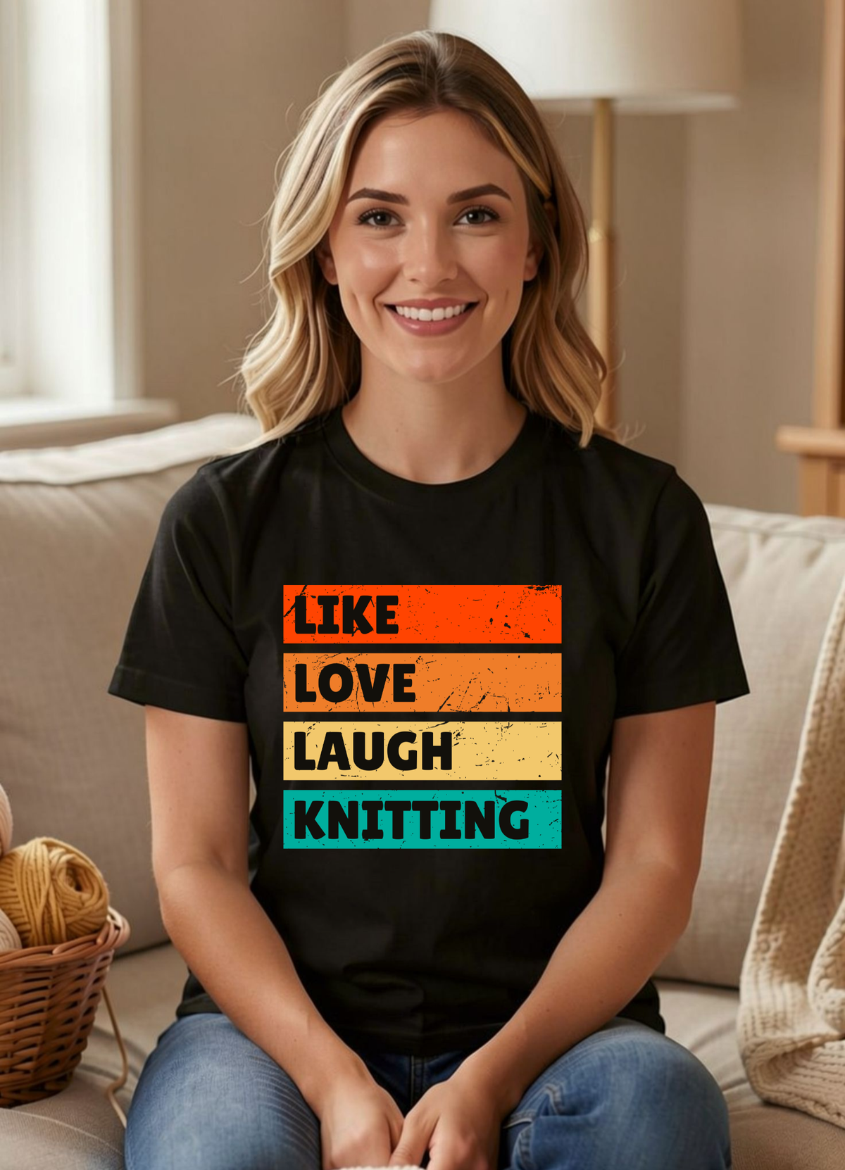 A smiling blonde woman sits on a beige couch wearing a black graphic T-shirt with stacked retro color blocks reading “LIKE LOVE LAUGH KNITTING” in orange, cream, and teal. A basket of yarn and home decor are visible in the softly lit living room setting.