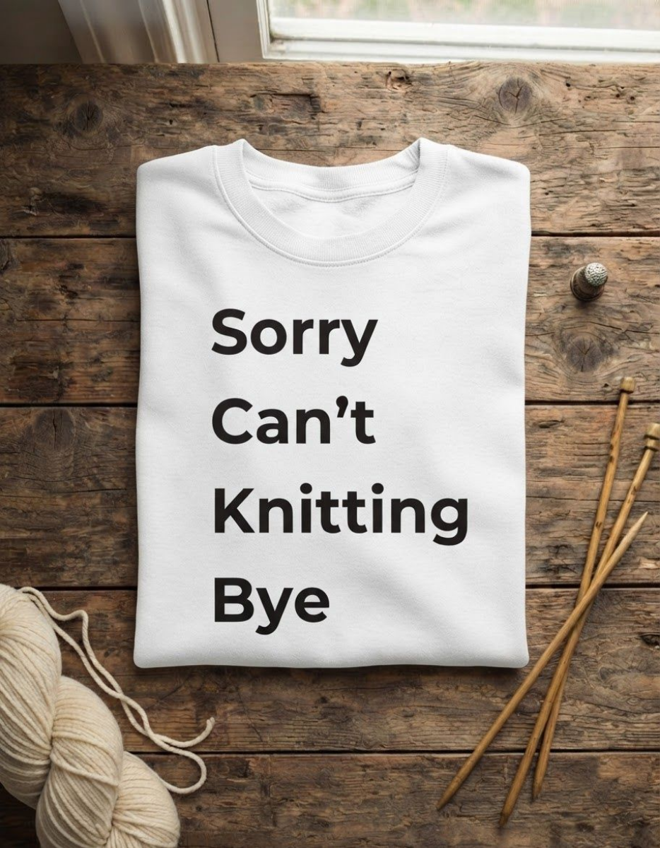 A folded white crewneck sweatshirt with the text 'Sorry Can't Knitting Bye' printed in black on the front, styled on a rustic wooden surface with knitting needles and yarn.