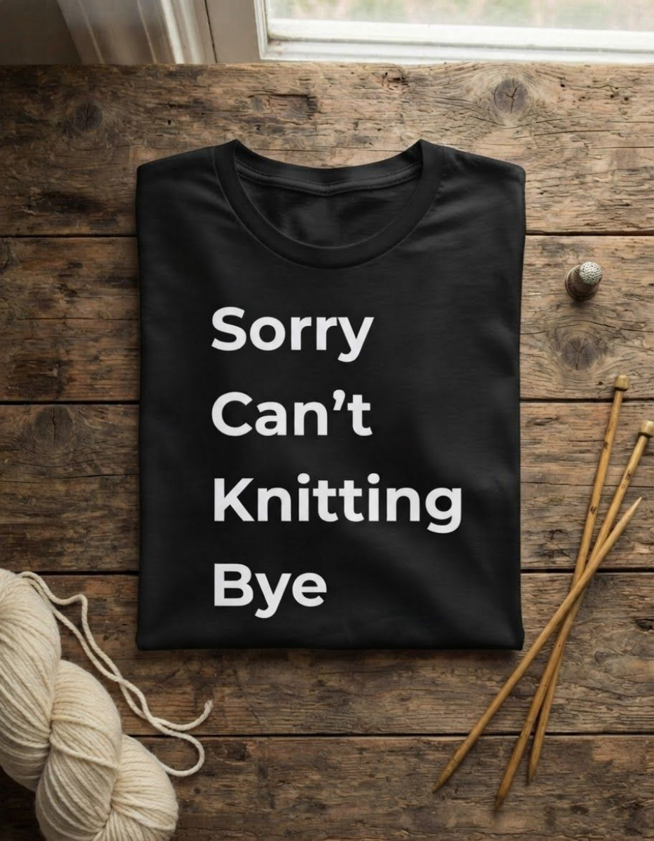 A black crewneck t-shirt with the text 'Sorry Can't Knitting Bye' printed in white, laid flat on a rustic wooden surface with knitting props including yarn and needles.