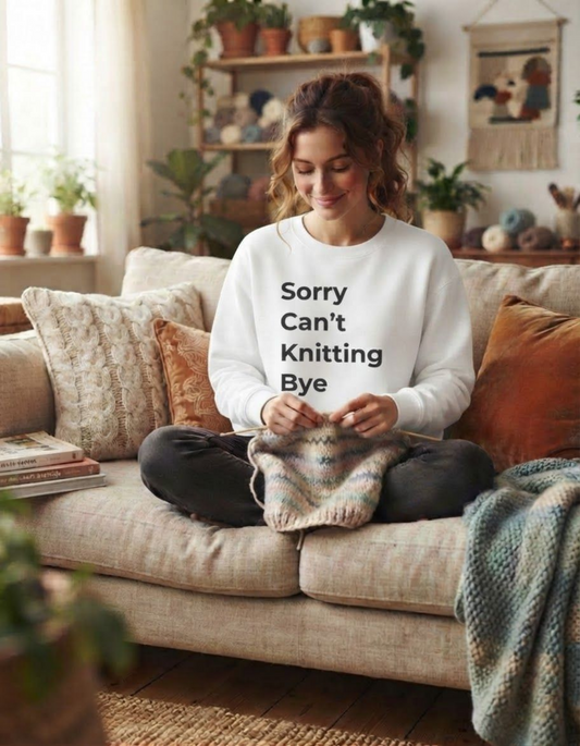 A woman sits cross-legged on a beige sofa in a cozy living room, knitting a small piece while wearing a white sweatshirt with the text 'Sorry Can't Knitting Bye' in black. The room has warm lighting, plants, shelves, and knitted textiles.