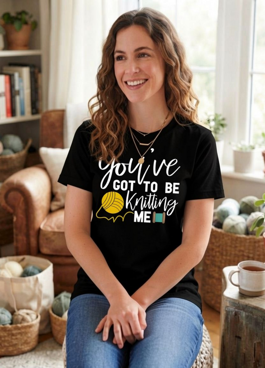 A smiling woman with wavy brown hair sits in a cozy living room setting, wearing a black graphic t-shirt that reads 'you've GOT TO BE Knitting ME' in white and yellow lettering with a yellow ball graphic. She is wearing blue jeans.