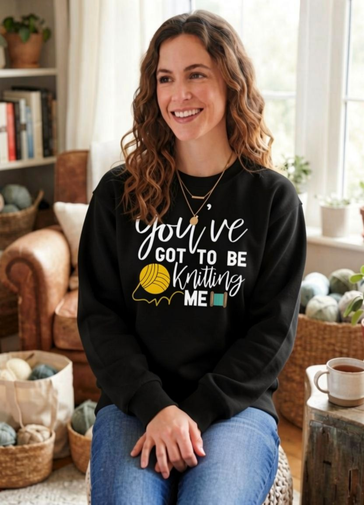 A smiling woman with wavy brown hair sits in a cozy living room setting wearing a black crewneck sweatshirt with a white and yellow graphic that reads 'you've GOT TO BE Knitting ME'. She is wearing blue jeans and sitting on a woven chair.