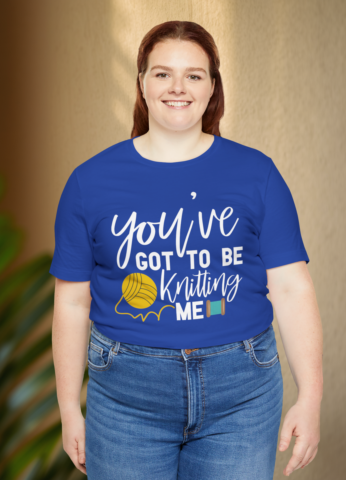 A woman wearing a royal blue graphic t-shirt with the text 'you've GOT TO BE Knitting ME' in white, accompanied by a yellow yarn ball graphic, paired with blue jeans against a warm beige background.