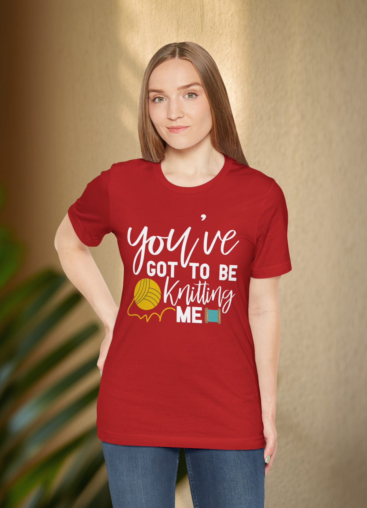 A woman modeling a red short-sleeve graphic t-shirt with the text 'you've GOT TO BE Knitting ME' in white script and illustration, paired with blue jeans against a warm beige textured wall background.